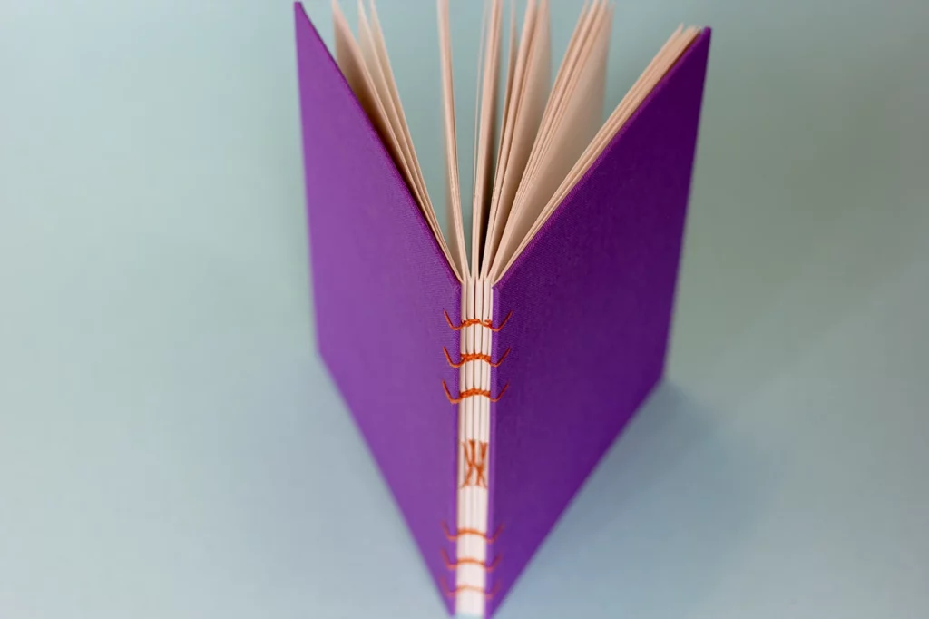 Carnet ouvert en reliure copte, couverture en toile du Marais violette et fil de lin orange, vue de dessus.