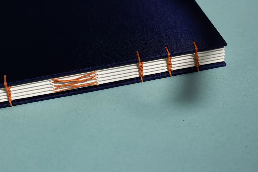 Zoom sur un carnet en reliure copte, révélant les chaînettes en fil de lin orange, couverture en toile bleue, réalisé dans mon atelier à Bordeaux.