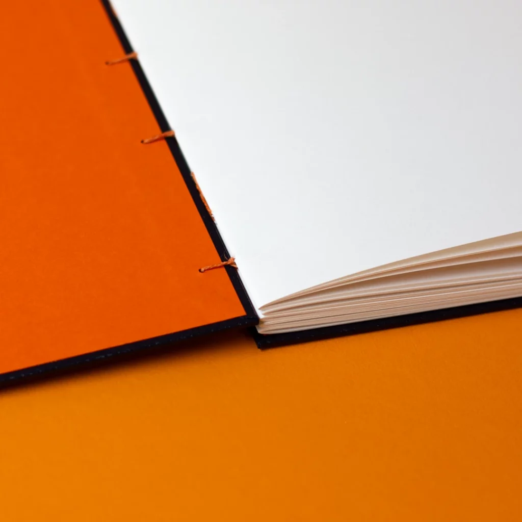 Carnet sur mesure ouvert avec reliure copte, couverture similicuir noir et fil de lin abricot, page de garde orange et papier Fedrigoni — Atelier Forme contre forme, Bordeaux.